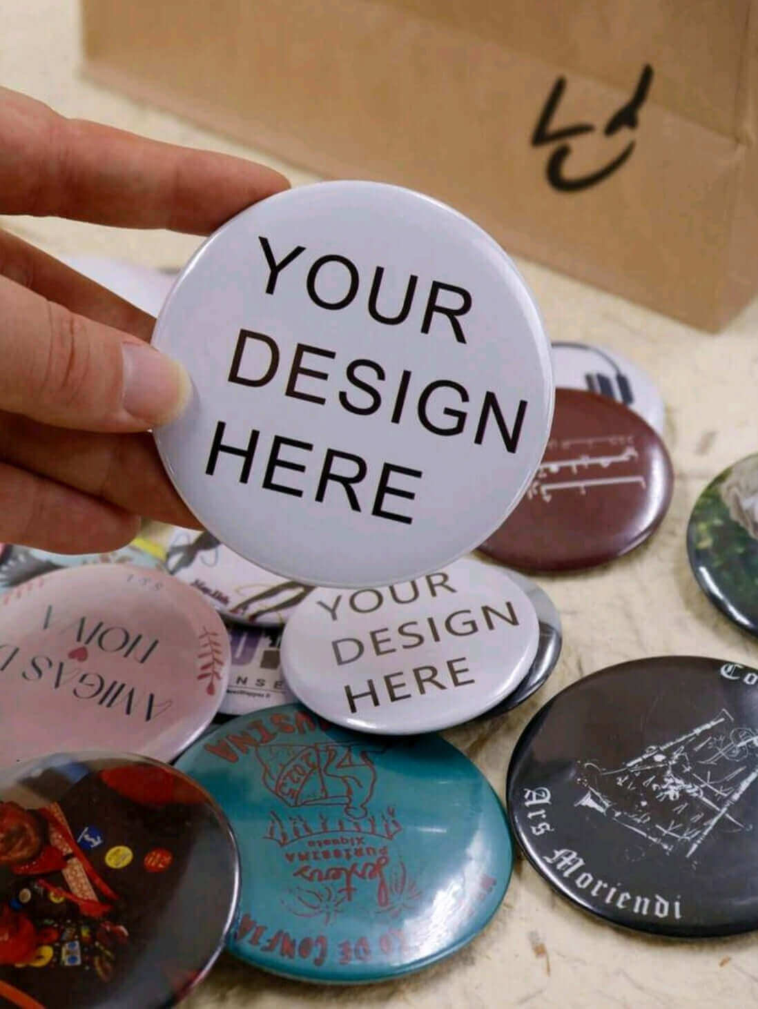 customised circle Pin with 'Your Design Here' text held by a hand, on a wooden surface.